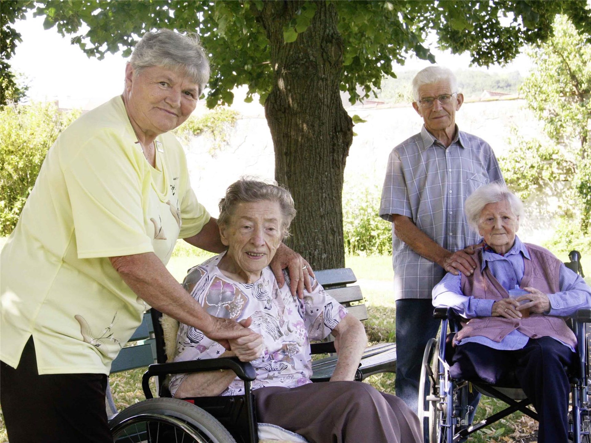 zwei ältere Personen kümmern sich um zwei noch ältere Personen in Rollstühlen in einem Garten