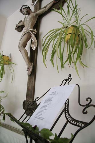 Ein Korpus am Kreuz an einer Wand hängend, daneben Blumenampeln, ebenfalls an der Wand hängend, davor ein Ständer mit einem Textblatt und Efeu 